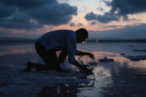 Vista lateral del hombre arrodillado y llenando el acuario vacío con agua del océano en la playa en el crepúsculo - foto de stock