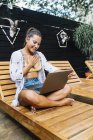 Mulher alegre em roupas casuais navegando laptop enquanto sentado com pernas cruzadas em cadeira de plataforma de madeira no terraço tropical — Fotografia de Stock