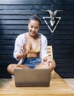 Donna allegra in abiti casual navigazione laptop mentre seduto con gambe incrociate sulla sedia a sdraio in legno sulla terrazza tropicale — Foto stock