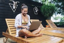 Mulher alegre em roupas casuais navegando laptop enquanto sentado com pernas cruzadas em cadeira de plataforma de madeira no terraço tropical — Fotografia de Stock