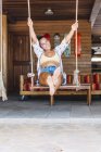 Braun gebrannte, glückliche Frau in Freizeitkleidung, die auf Schaukeln sitzt und wegschaut über eine Holzwand in Costa Rica — Stockfoto
