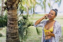 Счастливая женщина говорит на смартфоне, глядя в сторону, держа кружку и стоя на солнечной лужайке у пальмы в Коста-Рике — стоковое фото