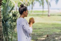 Vue de côté femme paisible buvant du café de tasse et debout près du palmier sur la mer ensoleillée au Costa Rica — Photo de stock