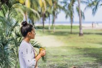 Vista lateral mujer pacífica beber café de taza y de pie junto a la palmera en la playa soleada en Costa Rica - foto de stock
