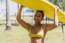 Mulher ajuste positivo em maiô carregando paddleboard amarelo sobre a cabeça na praia ensolarada por palmeiras na Costa Rica — Fotografia de Stock