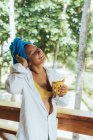 Zufriedene Frau in Kopftuch genießt Heißgetränk, während sie an einem Holzgeländer steht und auf grüne Bäume in Costa Rica blickt — Stockfoto