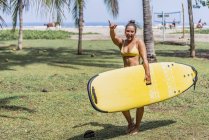 Positive fit woman in swimsuit holding yellow paddleboard and standing in sunny seashore by palm trees in Costa Rica — Stock Photo