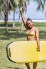 Позитивна жінка в купальнику, що тримає жовтий весло-борт і стоїть на сонячному узбережжі біля пальм у Коста-Риці. — стокове фото