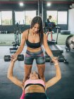 Smiling fit woman in sportswear insuring strong female partner lying on fitness bench and working out with weights — Stock Photo