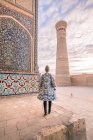 Visão traseira da fêmea em casaco ornamental em pé na praça envelhecida contra o céu nublado em Bucara, Uzbequistão — Fotografia de Stock