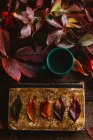 Сверху старинная книга с ярко-красными оранжево-желтыми осенними листьями и зеленой чашкой с напитком на деревянном столе — стоковое фото