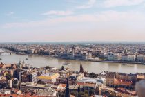 Лодки на спокойной реке, отражающей яркое небо и протекающей под мостом вдоль густонаселенного города Будапешта — стоковое фото