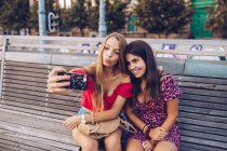 Mulheres rindo tomando selfie no banco de madeira na rua — Fotografia de Stock