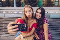 Riendo mujeres tomando selfie en banco de madera en la calle - foto de stock
