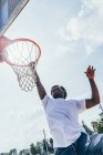 Poderoso deportista afroamericano enérgico colgado en la vuelta de baloncesto después de anotar la pelota en la red en el patio de recreo - foto de stock