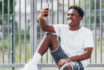 Pensivo sportivo afroamericano che naviga sul telefono cellulare sulla recinzione del parco giochi nella giornata luminosa — Foto stock