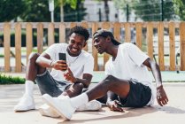 Cheerful amusing African American sportsmen surfing mobile phone comfortable placed on playground in bright day — Stock Photo