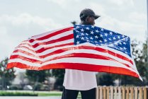 Hombre afroamericano serio sosteniendo la bandera americana en el hombro y mirando hacia otro lado - foto de stock