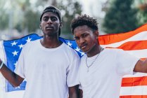 Hombres afroamericanos serios sosteniendo la bandera americana en el hombro y mirando a la cámara - foto de stock