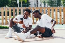 Alegre divertidos deportistas afroamericanos surfeando teléfono móvil cómodo colocado en el patio de recreo en día brillante - foto de stock