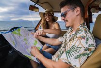 Side view of active smiling man in sunglasses looking at open road map and finding route with woman in hat in car — Stock Photo