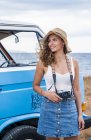 Обаятельная веселая дама в шляпе, держа камеру в руках рядом синий автомобиль на пляже и оглядываясь по сторонам — стоковое фото