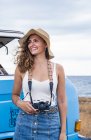Charmante dame joyeuse en chapeau tenant caméra à proximité voiture bleue sur la plage et regardant autour — Photo de stock