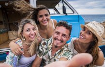 Laughing group of young people having fun taking selfie by blue minivan on beach in sunny daytime — Stock Photo