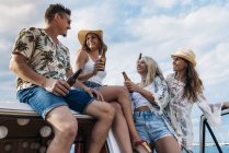 Cheerful lazy ladies with long hair in hats raising bottles of drinks and clinking with stylish man in car roof — Stock Photo