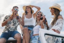 Cheerful lazy ladies with long hair in hats raising bottles of drinks and clinking with stylish man in car roof — Stock Photo