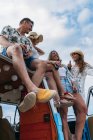 Grupo alegre de jovens e mulheres com garrafas de ligação sentado no telhado de minivan brilhante na praia durante o dia ensolarado — Fotografia de Stock