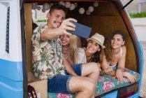 Pleasant group of young friends in trunk of minivan taking selfie on phone on beach in sunny daytime — Stock Photo