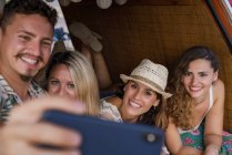 Agréable groupe de jeunes amis dans le coffre de minifourgonnette prendre selfie sur le téléphone sur la plage en plein jour ensoleillé — Photo de stock