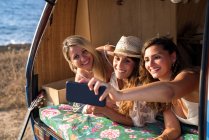 Веселые приятные дамы в багажнике яркого минивэна веселятся, делая селфи на мобильном телефоне на пляже в солнечный день — стоковое фото