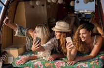 Senhoras alegres e agradáveis deitadas no porta-malas da minivan brilhante e se divertindo tirando selfies em telefones celulares na praia — Fotografia de Stock