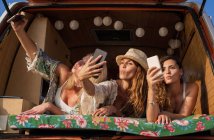 Cheerful pleasant ladies lying on trunk of bright minivan and having fun taking selfies on mobile phones on beach — Stock Photo