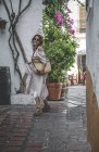 Vista lateral de la mujer entusiasta positiva de moda con bolsa de verano disfrutando paseando por la calle trasera de Marbella - foto de stock