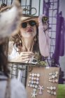Стильна впевнена жінка в сонцезахисних окулярах дивиться в дзеркало для перевірки солом'яного капелюха з шнуром на міському ринку — стокове фото