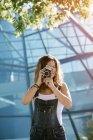 Joven mujer entusiasta capturando momento tomando fotos en la cámara en el fondo de la arquitectura de vidrio - foto de stock