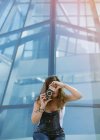 Молодая увлеченная женщина снимает момент на камеру на фоне стеклянной архитектуры — стоковое фото