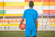 Вид сзади на черного парня в синей спортивной форме, несущего красный мяч, стоя напротив стадиона на футбольном поле — стоковое фото