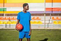 Jogador de futebol preto com bola em pé no estádio e olhando para longe — Fotografia de Stock