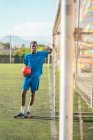 Negro adolescente mirando en cámara como inclinado en gol post en campo de fútbol - foto de stock