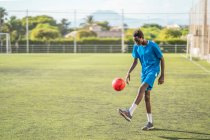 Adolescente étnico em azul sportswear malabarismo bola vermelha durante o treino no campo de futebol — Fotografia de Stock