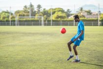 Adolescent ethnique en vêtements de sport bleus jonglant avec la balle rouge pendant l'entraînement sur le terrain de football — Photo de stock