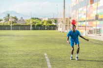 Flaco negro adolescente jugando pelota de fútbol en la cabeza en el campo verde - foto de stock