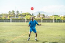 Flaco negro adolescente jugando pelota de fútbol en la cabeza en el campo verde - foto de stock