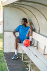 Повна довжина афроамериканського підлітка з футбольним м'ячем сидить на лавці і озирається під час тренувань — стокове фото