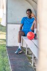 Full length of African American teenager with football ball sitting on bench and looking in camera during workout — Stock Photo
