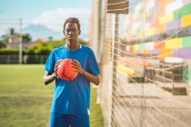 Черный подросток смотрит в камеру с красным мячом в руках на футбольном поле — стоковое фото
